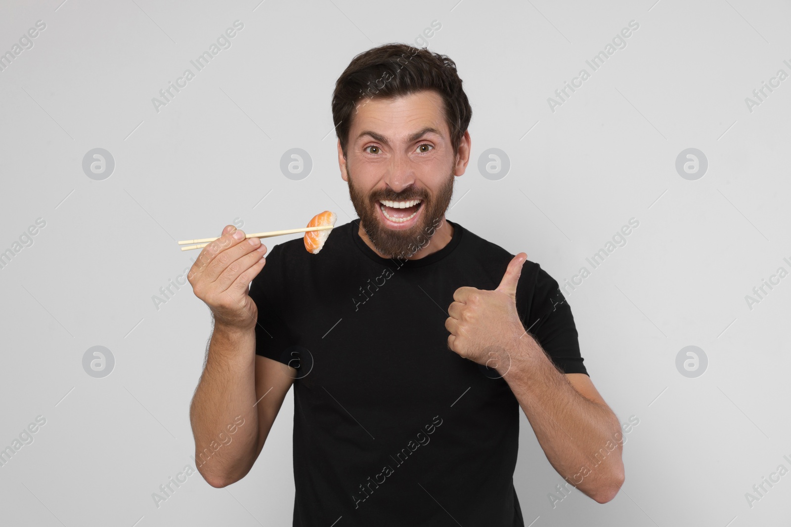 Photo of Emotional man holding tasty sushi with chopsticks and showing thumbs up on light grey background