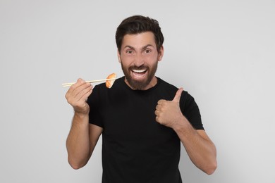 Photo of Emotional man holding tasty sushi with chopsticks and showing thumbs up on light grey background