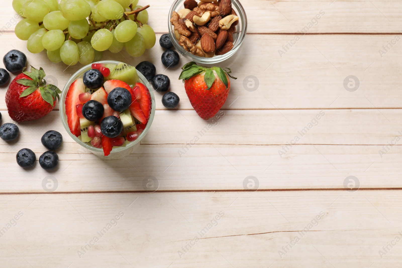 Photo of Delicious fruit salad in glass, nuts and ingredients on light wooden table, flat lay. Space for text