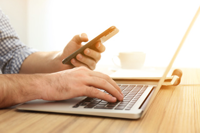 Man with smartphone working on laptop at wooden table indoors, closeup