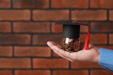 Photo of Man holding glass jar of coins and graduation cap against brick background, closeup with space for text. Scholarship concept