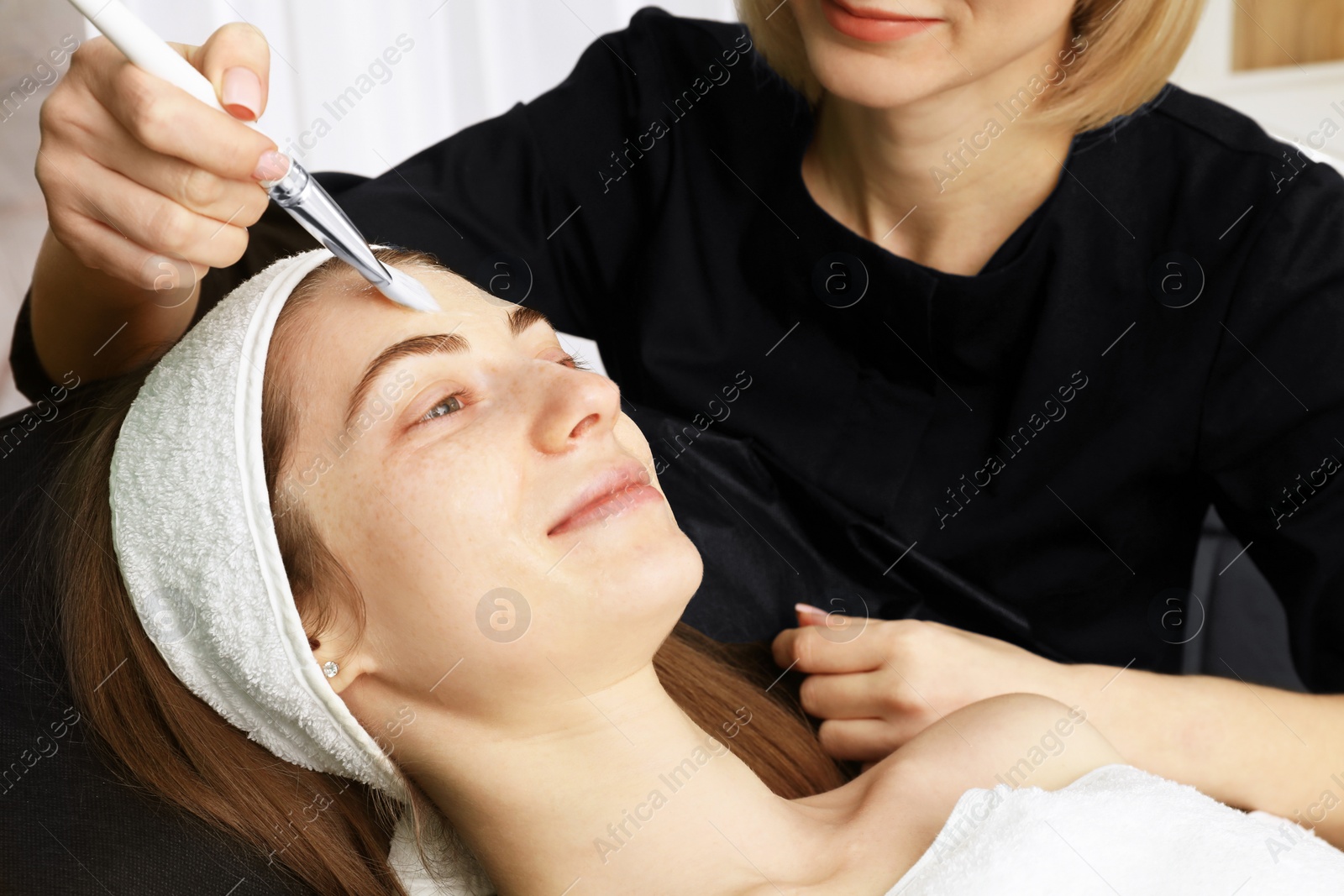 Photo of Cosmetologist applying mask on woman's face indoors, closeup