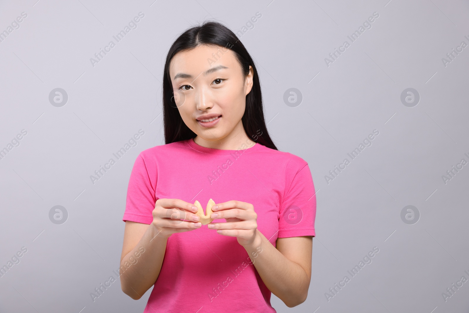 Photo of Asian woman holding tasty fortune cookie with prediction on grey background, space for text