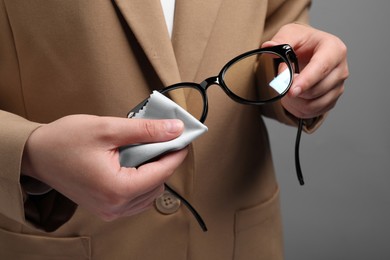 Woman wiping her glasses with microfiber cloth on grey background, closeup