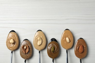 Photo of Tasty nut butters in spoons and raw nuts on white wooden table, flat lay. Space for text