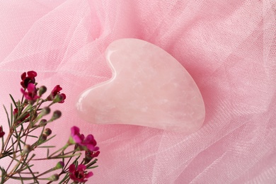 Photo of Rose quartz gua sha tool and flowers on pink fabric, flat lay