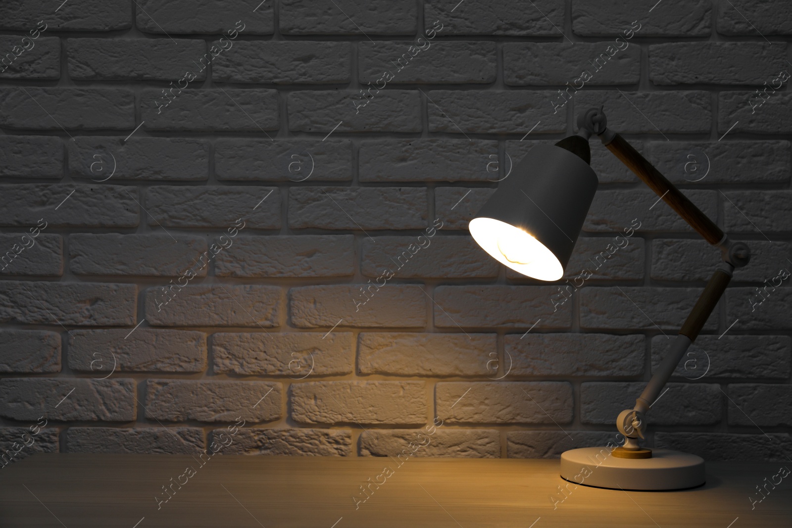 Photo of Stylish modern desk lamp on white wooden table near brick wall at night, space for text