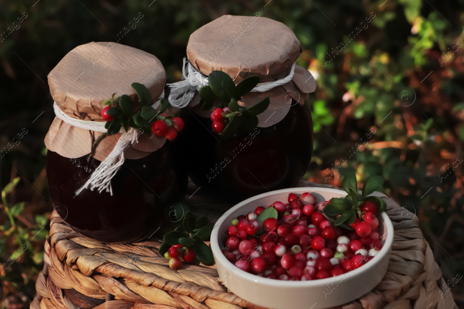 Photo of Jars of delicious lingonberry jam and red berries on wicker basket outdoors