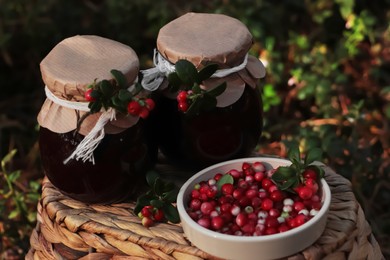 Jars of delicious lingonberry jam and red berries on wicker basket outdoors