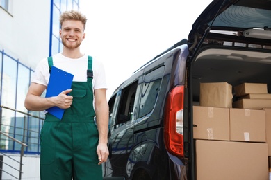 Photo of Young courier holding clipboard near delivery van with parcels outdoors