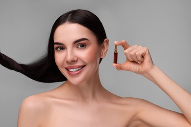 Beautiful young woman with long healthy hair holding ampoule on grey background