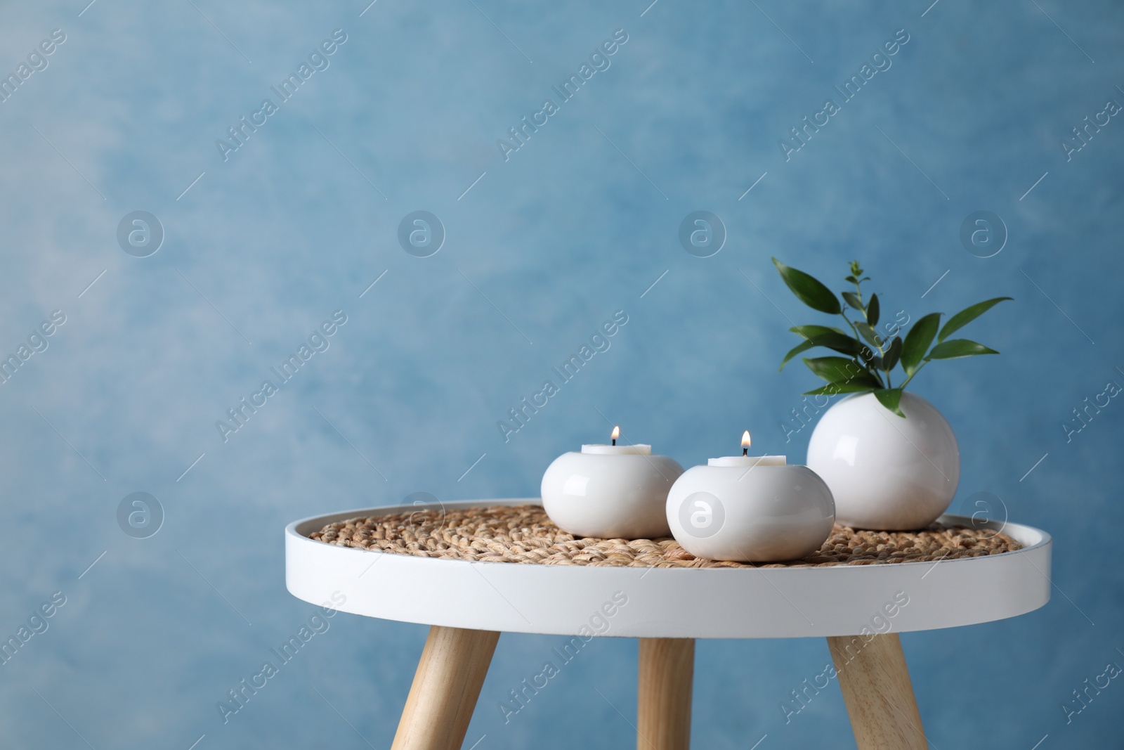 Photo of Burning candles and plant on white table against light blue background, space for text