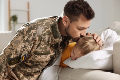 Photo of Soldier in Ukrainian military uniform kissing his daughter while she sleeping on sofa at home. Family reunion