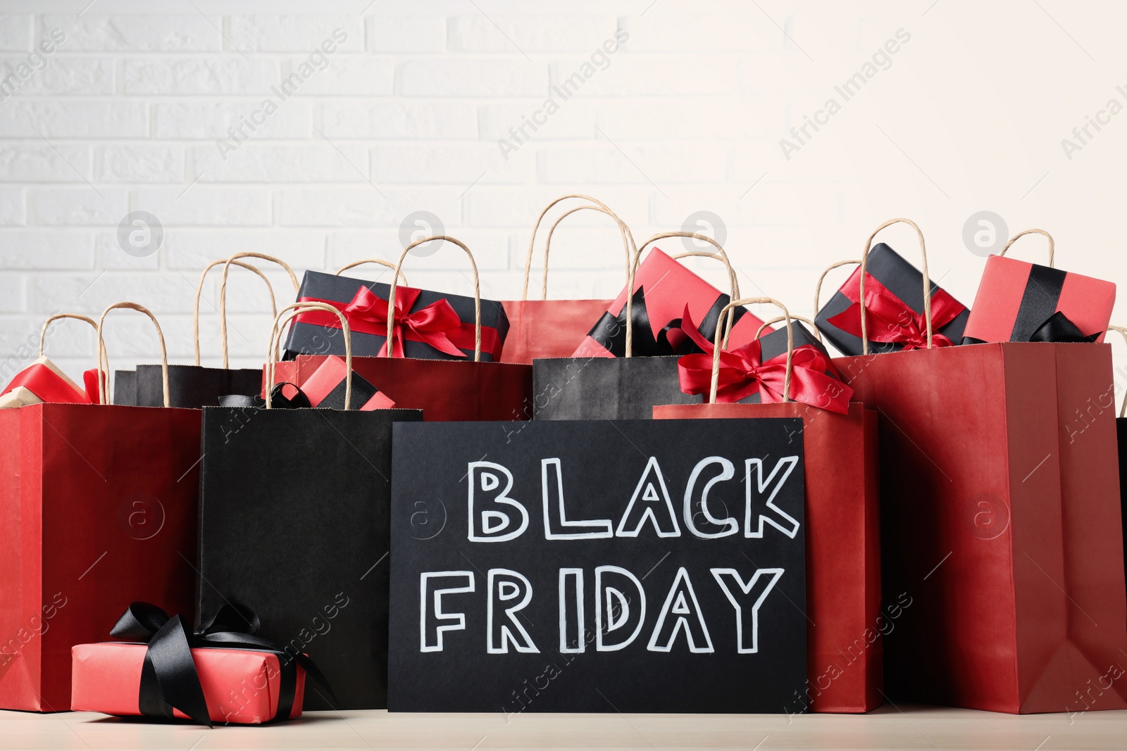 Photo of Many shopping bags, gift boxes and card with words Black Friday on table against white brick wall