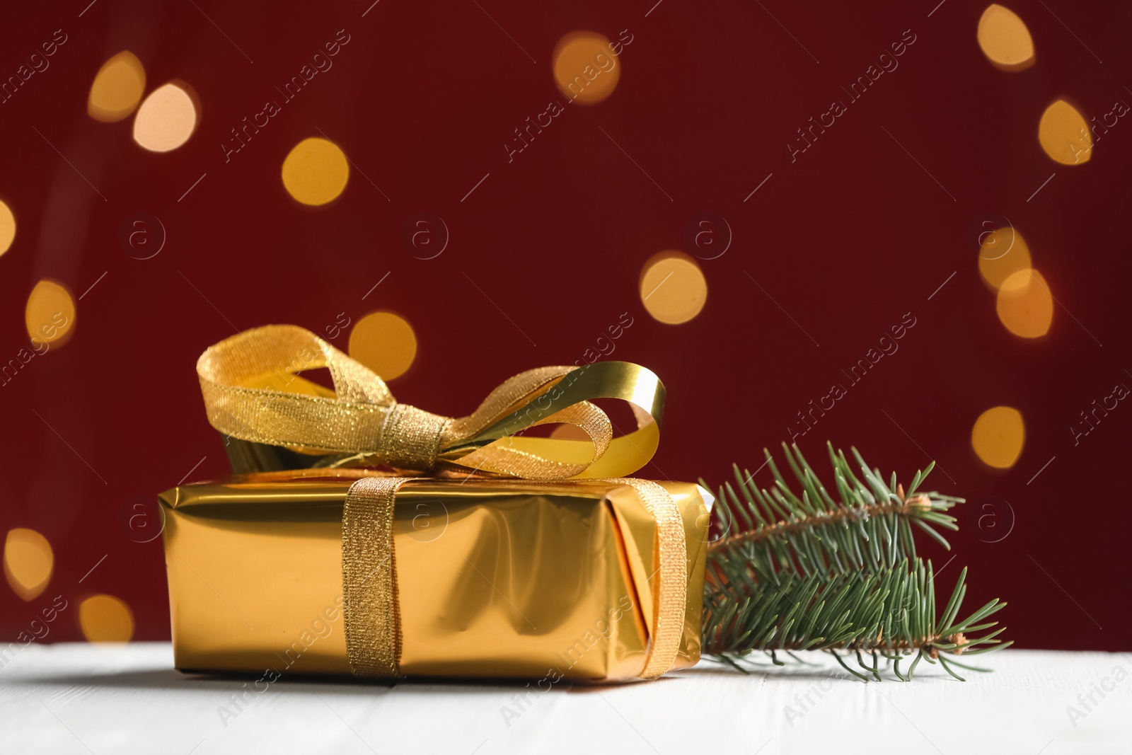Photo of Golden gift box and fir tree twig against red background with blurred lights, space for text