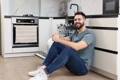 Photo of Smiling man sitting near open dishwasher in kitchen