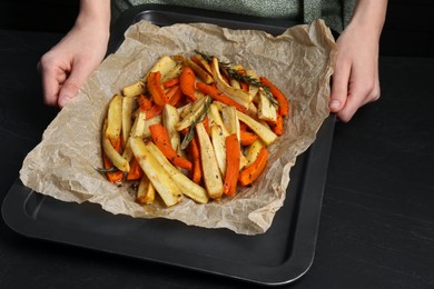 Photo of Cooker holding baking pan with tasty parsnip and bell pepper at black table, closeup