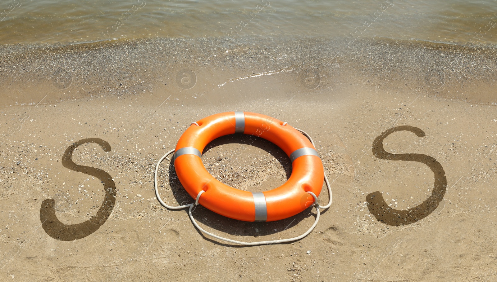 Image of SOS message handwritten on sandy beach with lifebuoy near sea