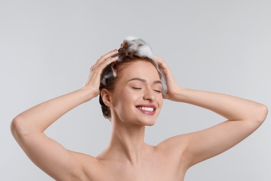 Happy young woman washing her hair with shampoo on light grey background