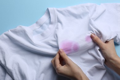 Woman holding white shirt with purple stains on light blue background, top view