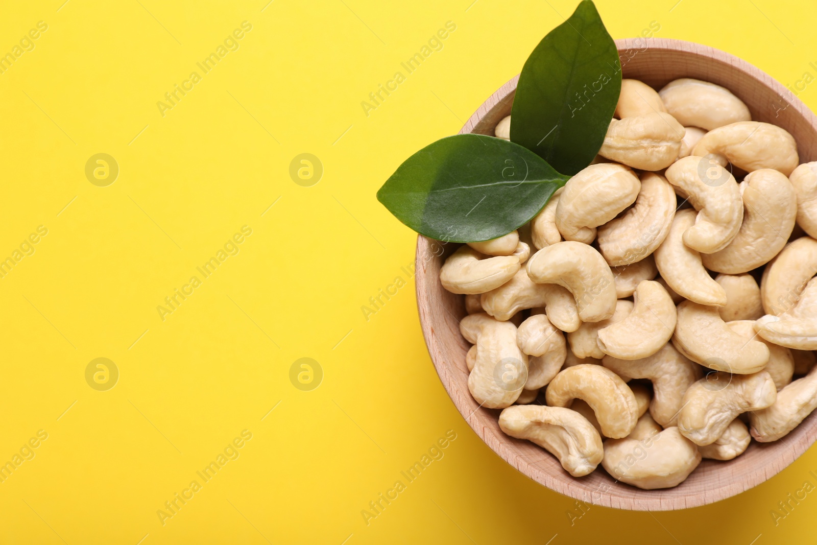 Photo of Tasty cashew nuts and green leaves in bowl on yellow background, top view. Space for text