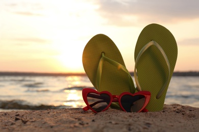 Stylish flip flops with sunglasses on sand near sea, space for text. Beach accessories