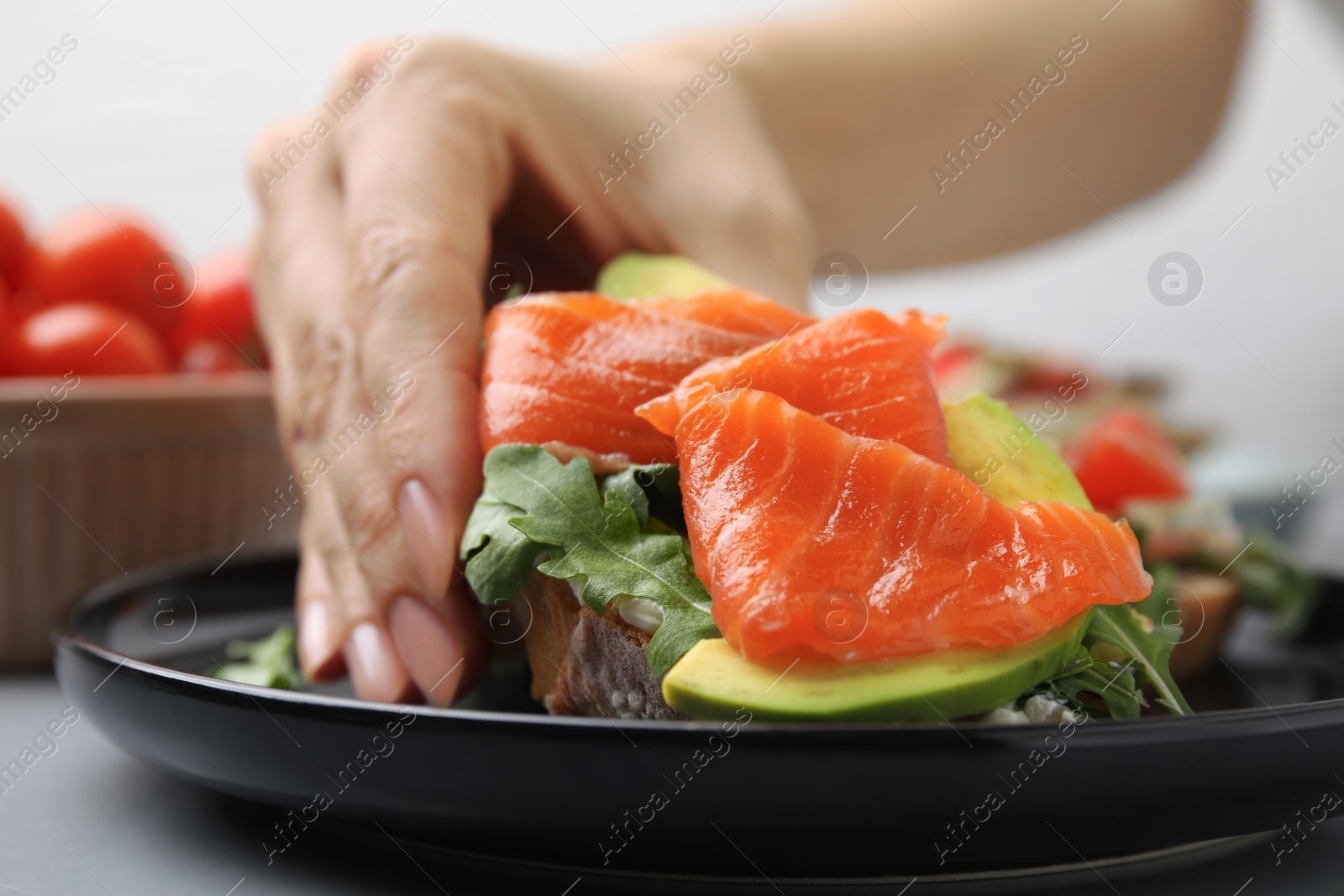 Photo of Woman taking tasty bruschetta with salmon, arugula and avocado from black plate, closeup