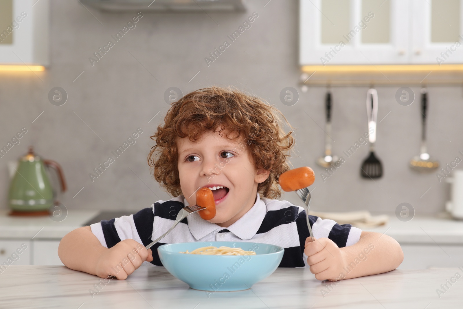 Photo of Cute little boy eating sausage and pasta at table in kitchen