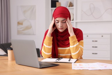 Photo of Tired Muslim woman working near laptop at wooden table in room