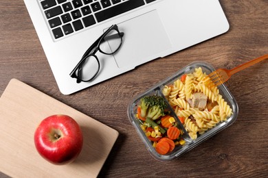 Container of tasty food, laptop, apple, book and glasses on wooden table, flat lay. Business lunch