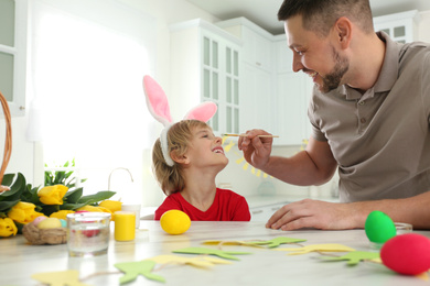 Happy son wearing bunny ears headband and his father having fun while at table in kitchen with Easter eggs