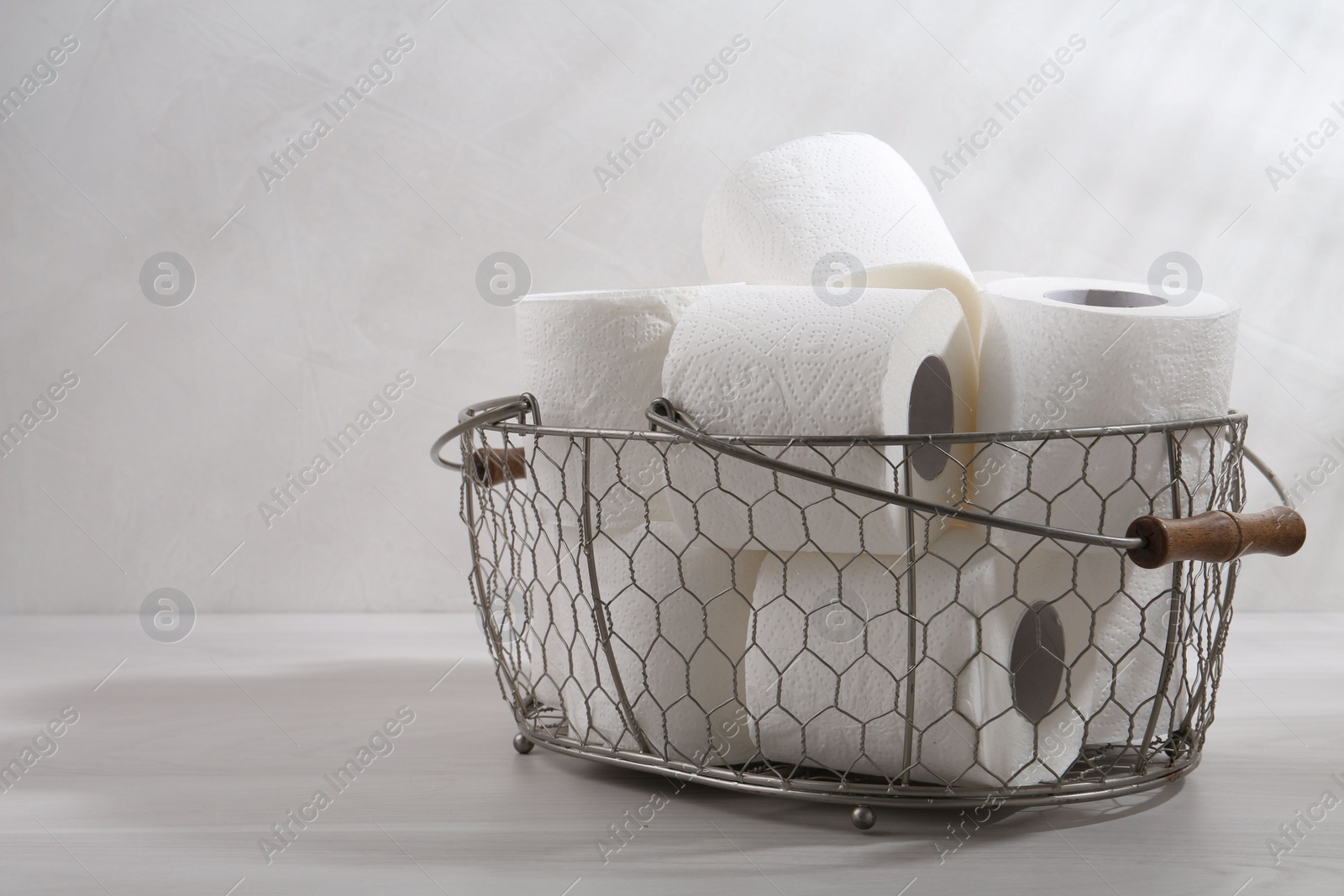 Photo of Soft toilet paper rolls in metal basket on white wooden table, closeup. Space for text