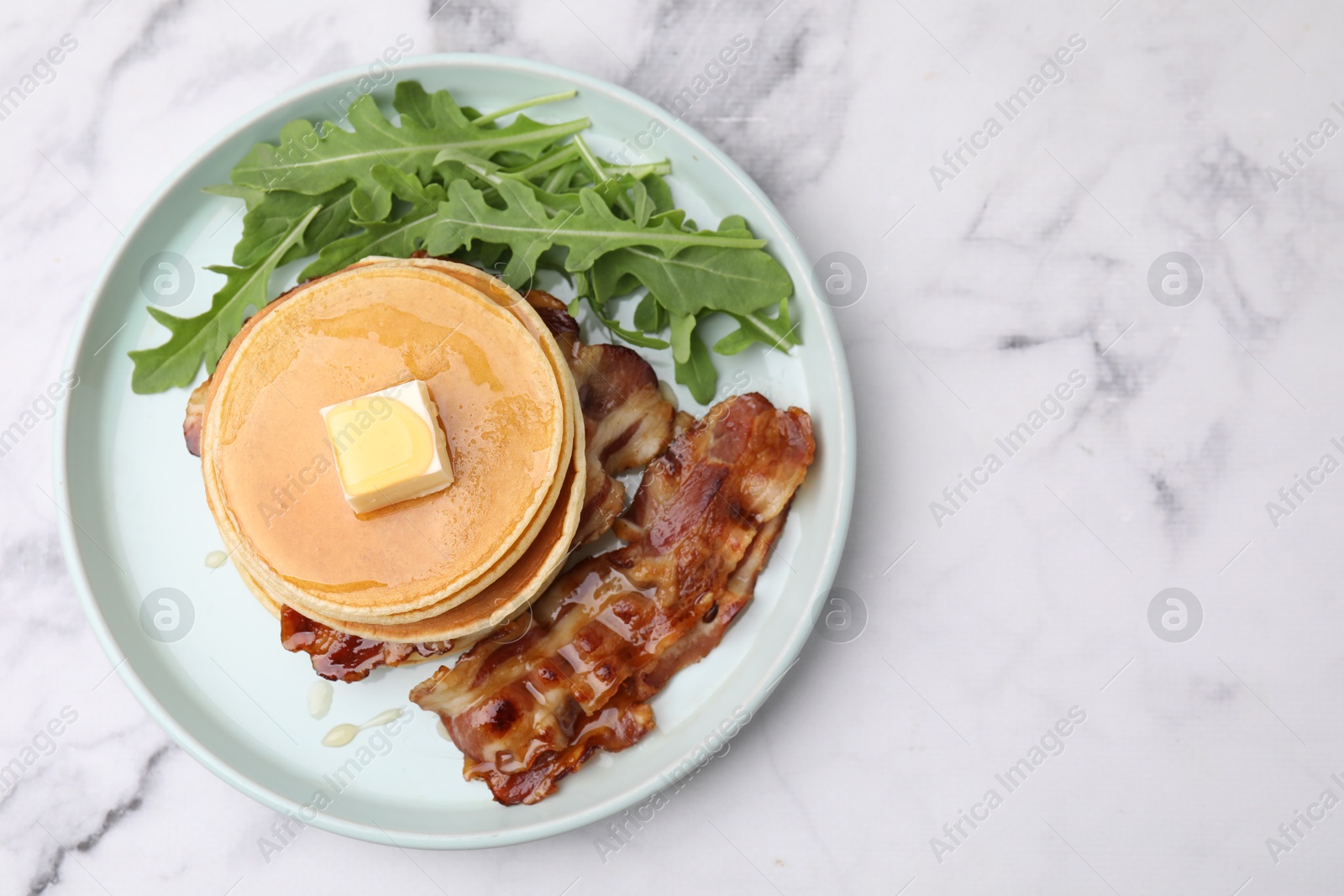 Photo of Tasty pancakes with butter, fried bacon and fresh arugula on white marble table, top view. Space for text