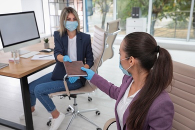 Photo of Office employee in mask and gloves giving notebook to her colleague at workplace