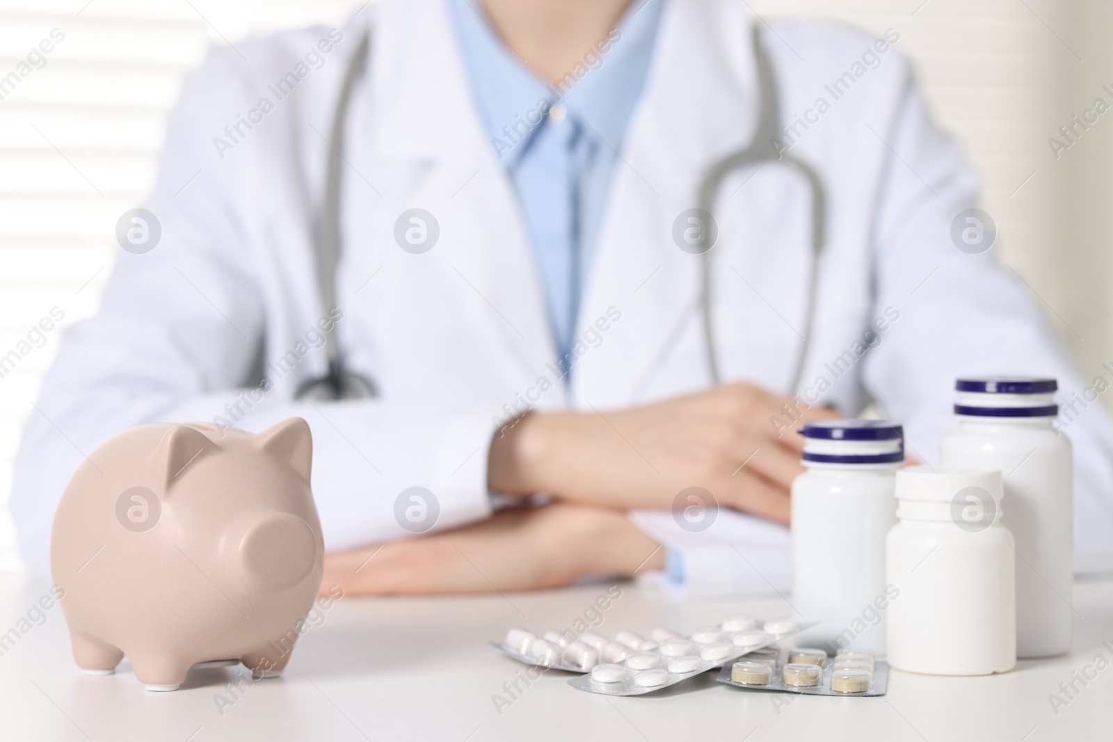 Photo of Doctor at white table indoors, focus on piggy bank and pills