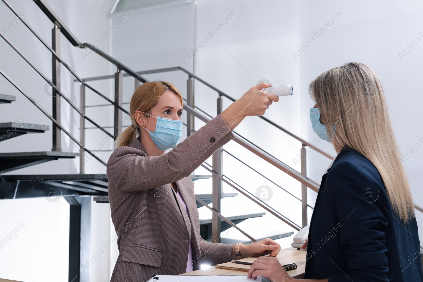 Photo of Woman in mask measuring temperature of employee with noncontact thermometer at office reception