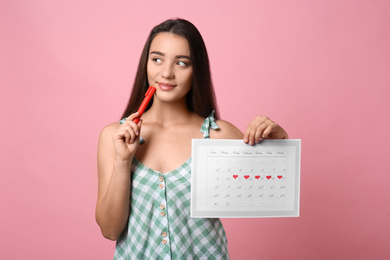 Young woman holding calendar with marked menstrual cycle days on pink background