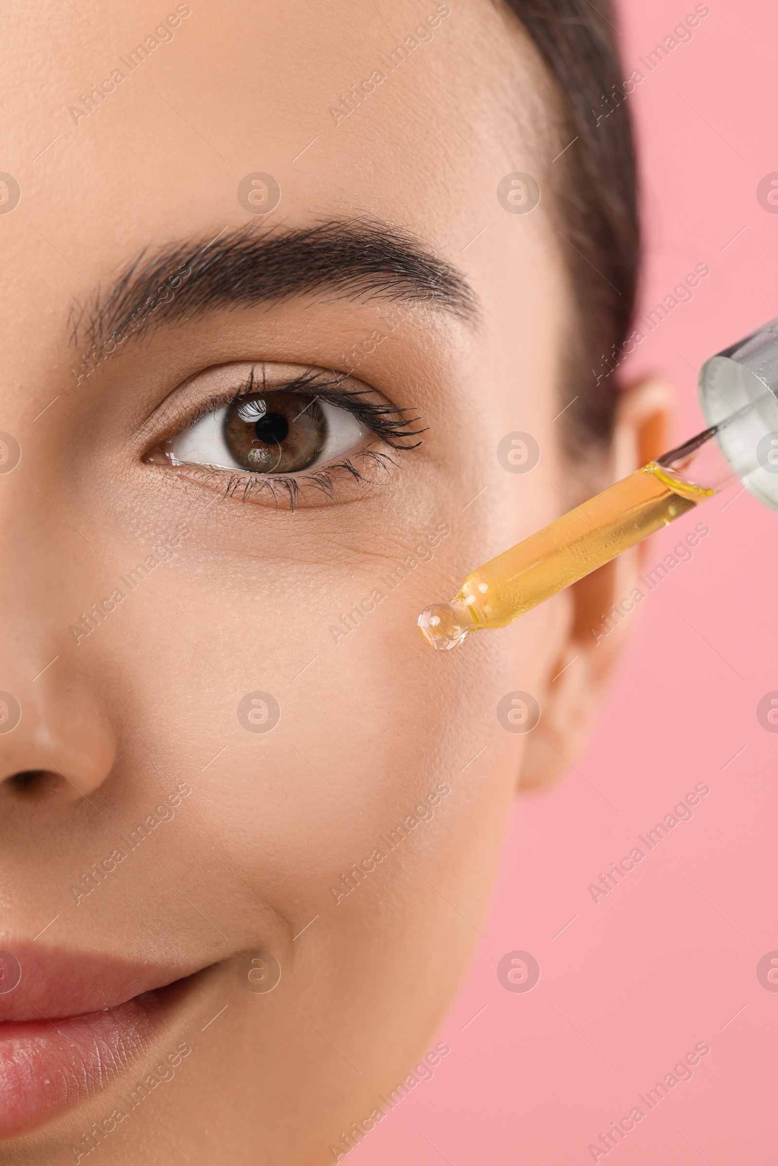 Photo of Beautiful young woman applying serum onto her face on pink background, closeup