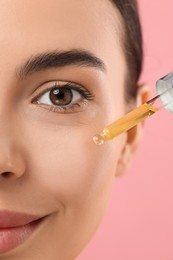 Beautiful young woman applying serum onto her face on pink background, closeup