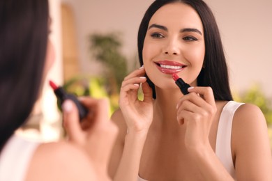 Photo of Young woman applying beautiful glossy lipstick in front of mirror indoors