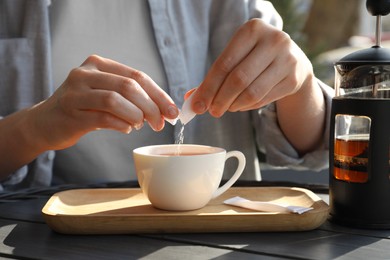 Woman adding sugar into cup of tea at black wooden table in outdoor cafe, closeup