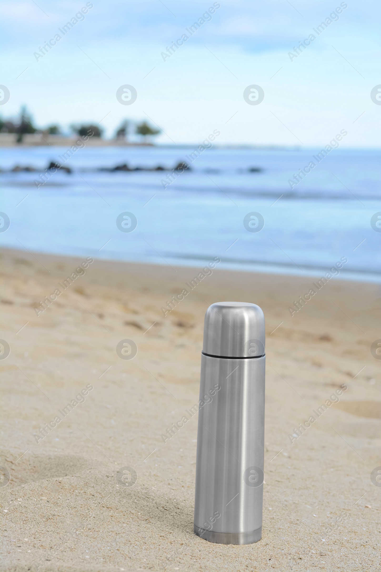 Photo of Metallic thermos with hot drink on sandy beach near sea, space for text