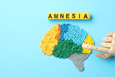 Photo of Word Amnesia, brain with sections made of plasticine and mannequin hand on light blue background, flat lay. Space for text