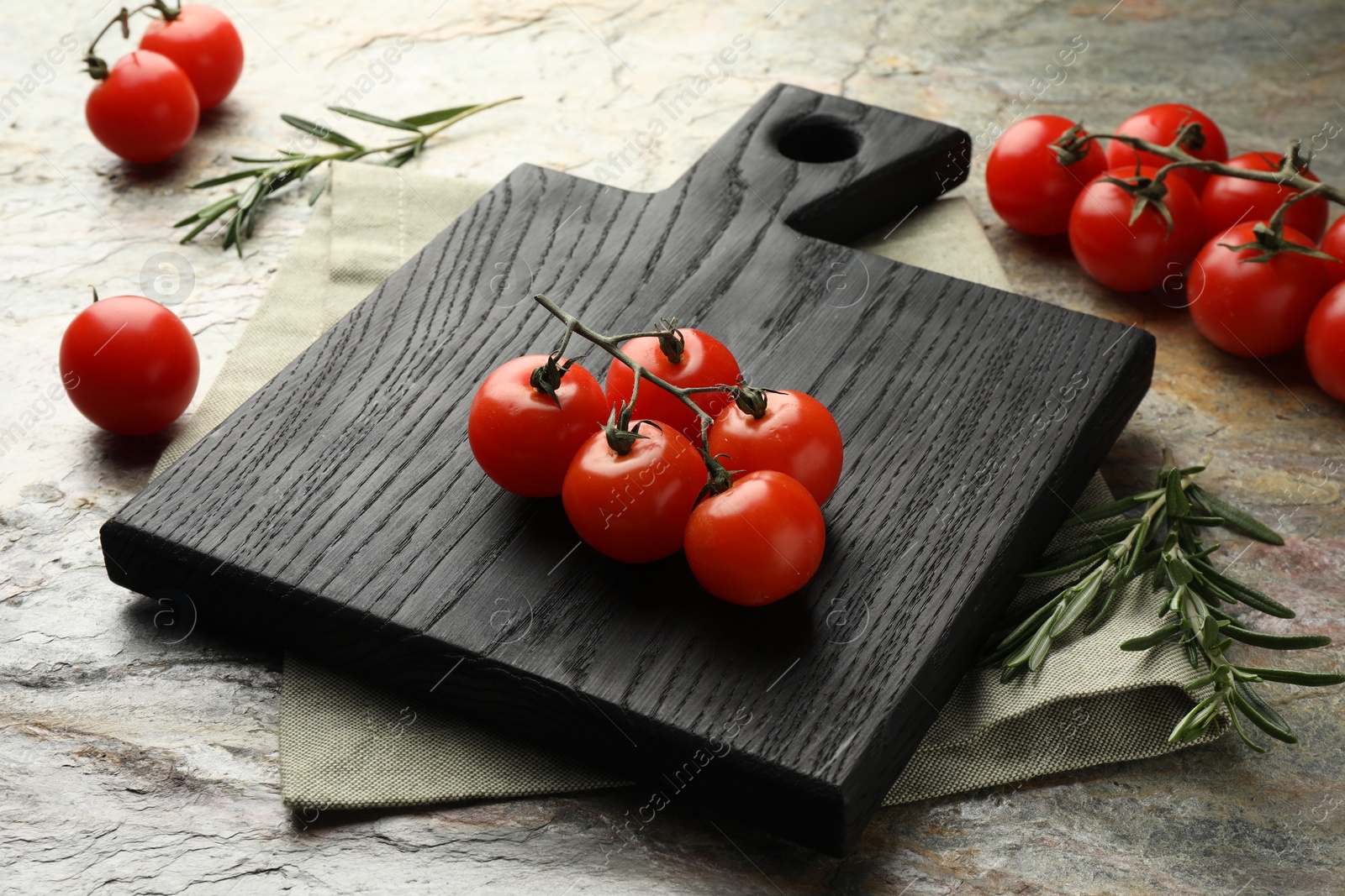 Photo of Black cutting board, tomatoes and rosemary on textured table, closeup