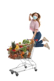 Photo of Young woman in medical mask with shopping cart full of groceries on white background