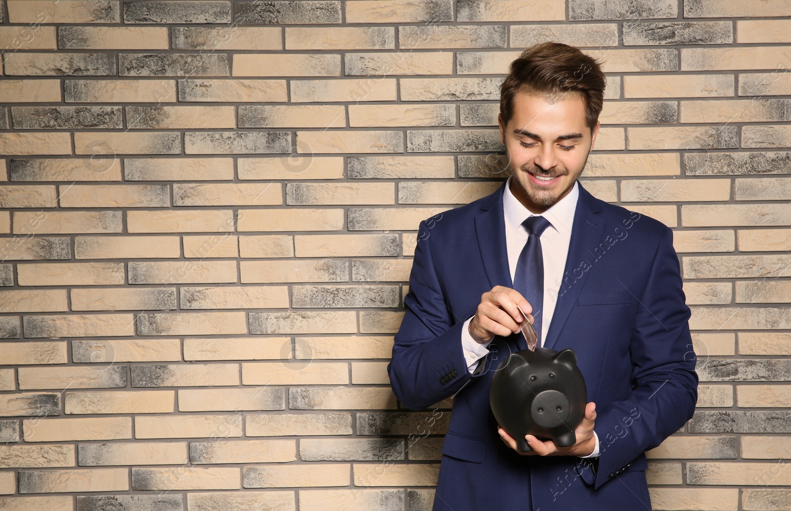 Photo of Young businessman putting money into piggy bank near brick wall. Space for text