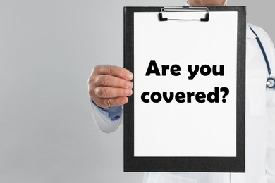 Image of Doctor holding clipboard with text ARE YOU COVERED? on light background, closeup. Insurance concept
