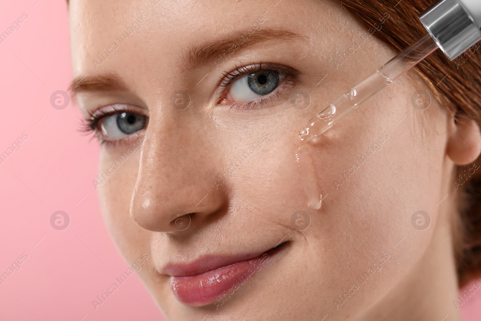 Photo of Beautiful woman with freckles applying cosmetic serum onto her face on pink background, closeup