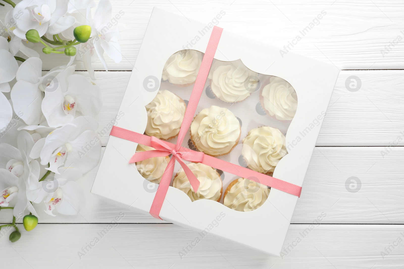 Photo of Tasty cupcakes with vanilla cream in box and orchid flowers on white wooden table, top view