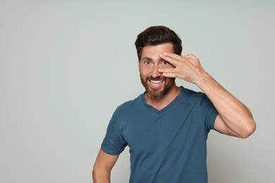 Smiling bearded man looking through spread fingers on grey background. Space for text